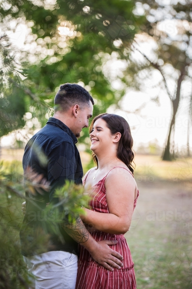 Boyfriend and girlfriend standing together behind tree looking and smiling at each other - Australian Stock Image