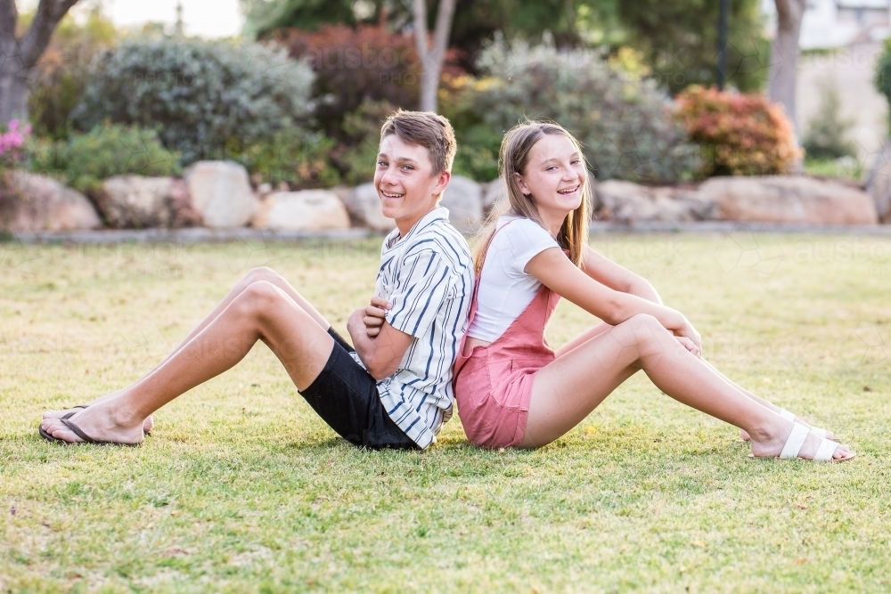 Brother and sister sitting back to back on grass in garden smiling - Australian Stock Image