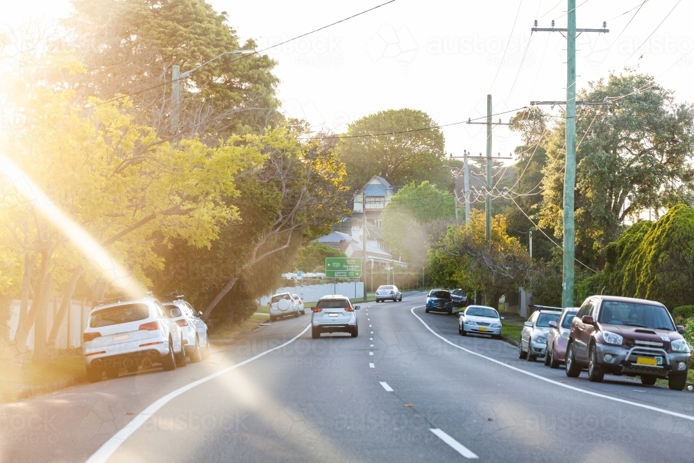 Car driving down road with parked cars beside in afternoon light - Australian Stock Image