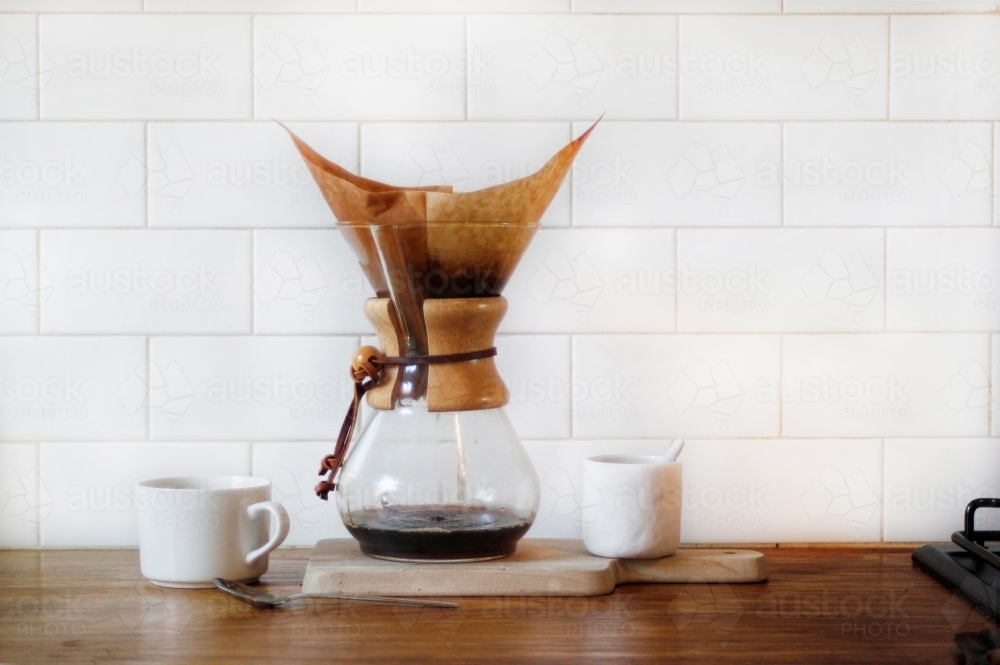 Chemex coffee filter on timber kitchen bench with coffee cup, sugar bowl against white subway tiles - Australian Stock Image