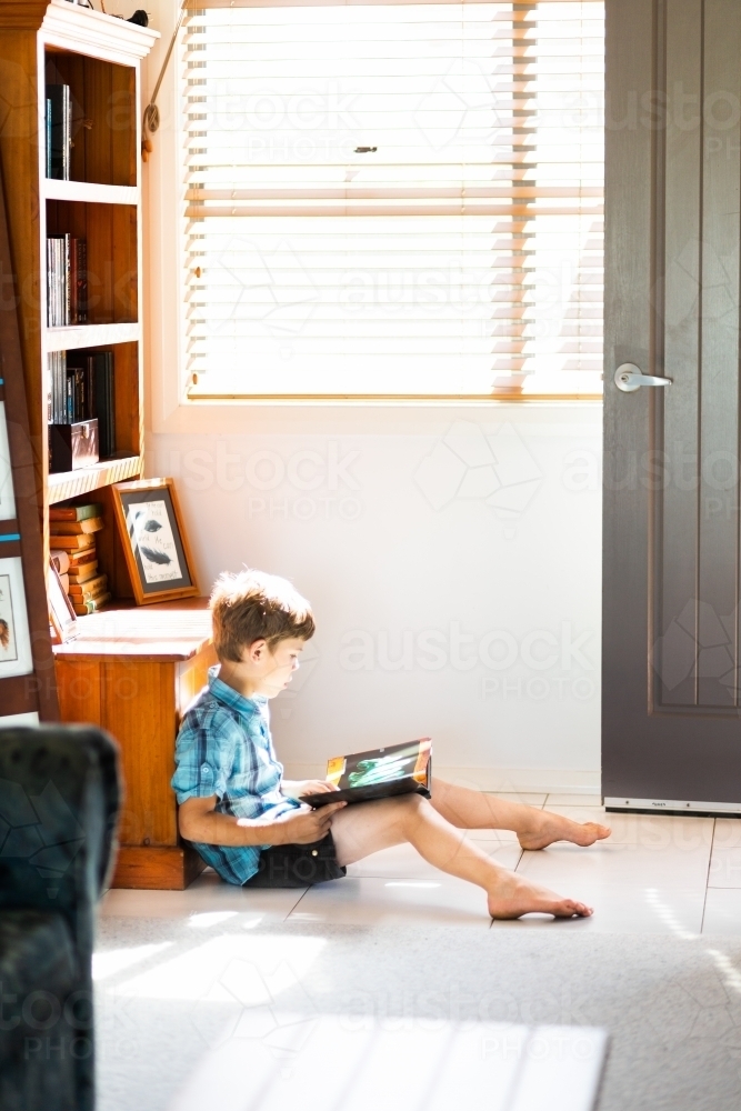 Child sitting in sunlight on the floor reading a book - Australian Stock Image