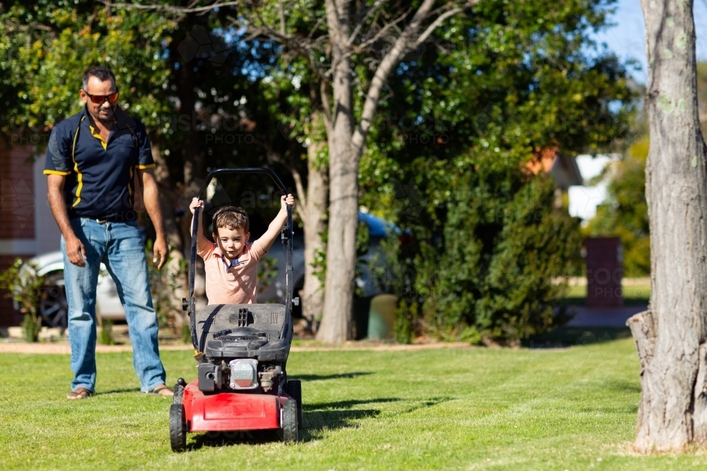 child trying to push lawnmower watched by father - Australian Stock Image