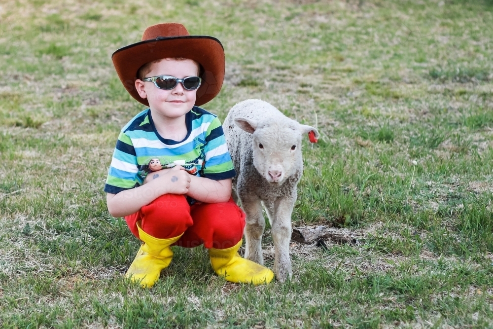 Child wearing akubra hat and sunglasses sitting with lamb - Australian Stock Image