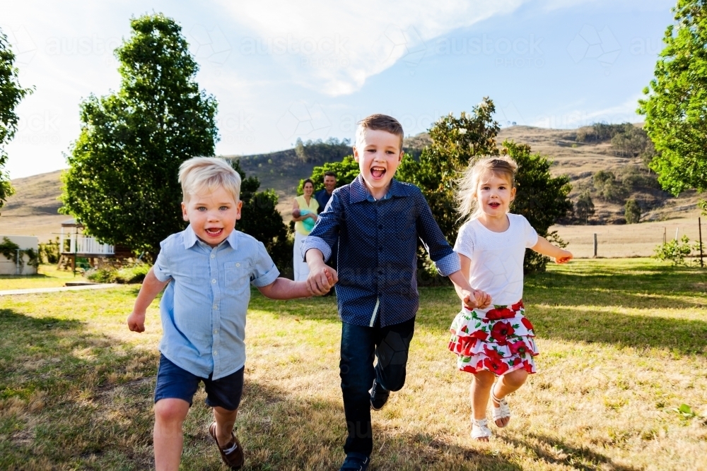 Children running through backyard holding hands with parents watching - Australian Stock Image