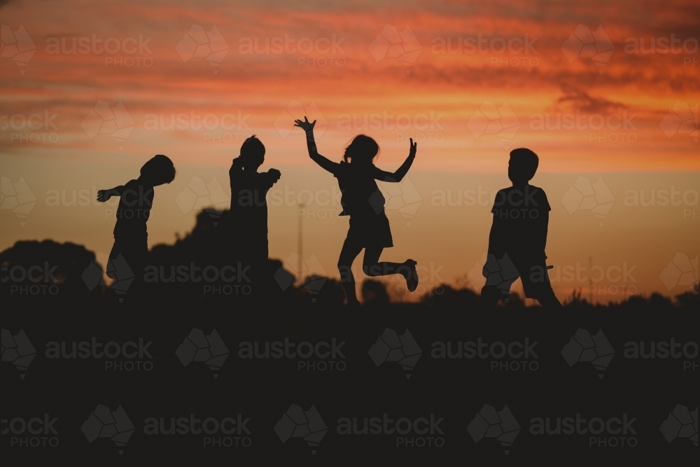 Children silhouette playing at sunset with girl jumping - Australian Stock Image