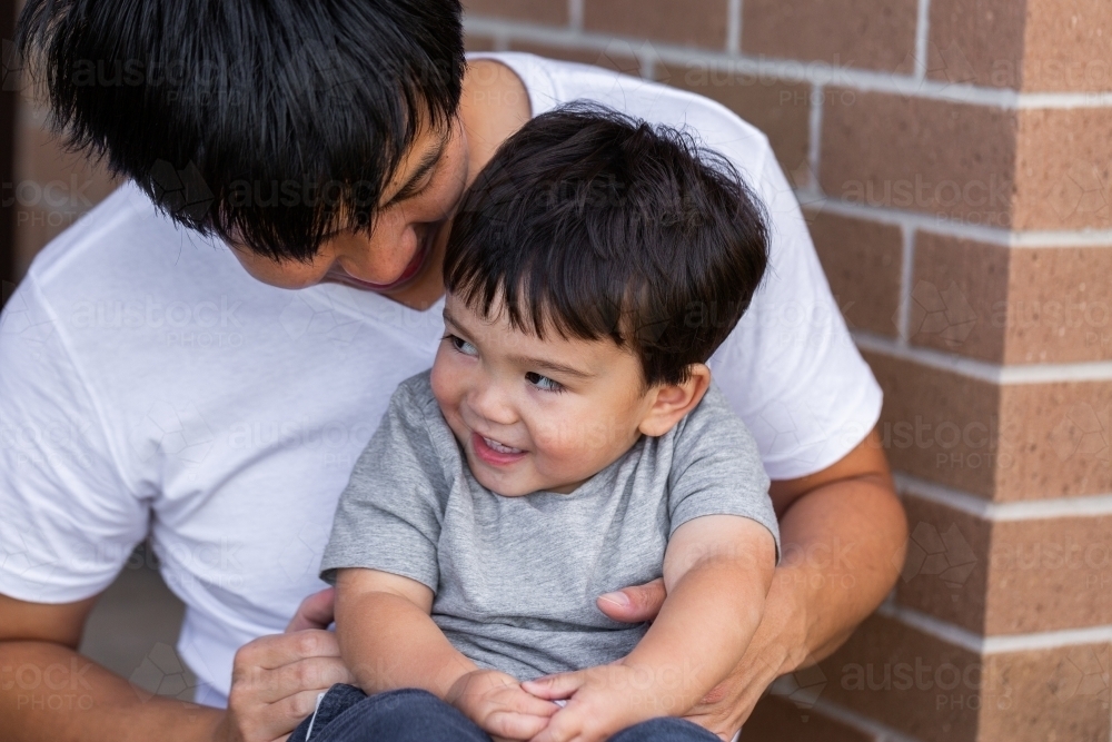 Chinese australian dad and toddler cuddle together by front of home - Australian Stock Image