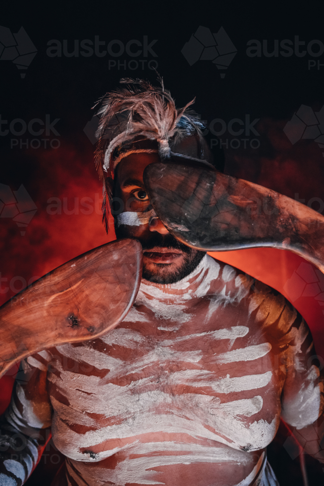 Close-up of aboriginal man holding a pair of boomerangs to his face. - Australian Stock Image