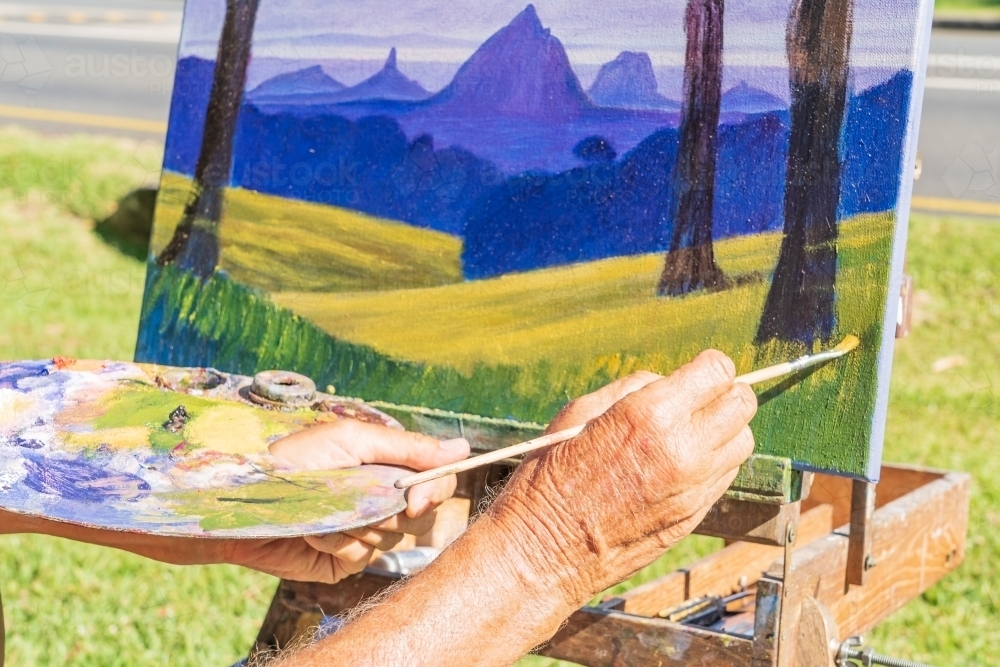 Close up of an artists hands painting a canvas and holding a palette - Australian Stock Image