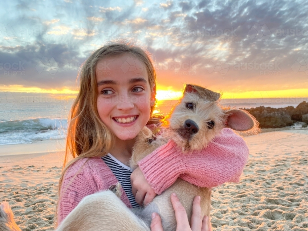 Close up of young girl cuddling puppy on the beach at sunset with ocean in background - Australian Stock Image