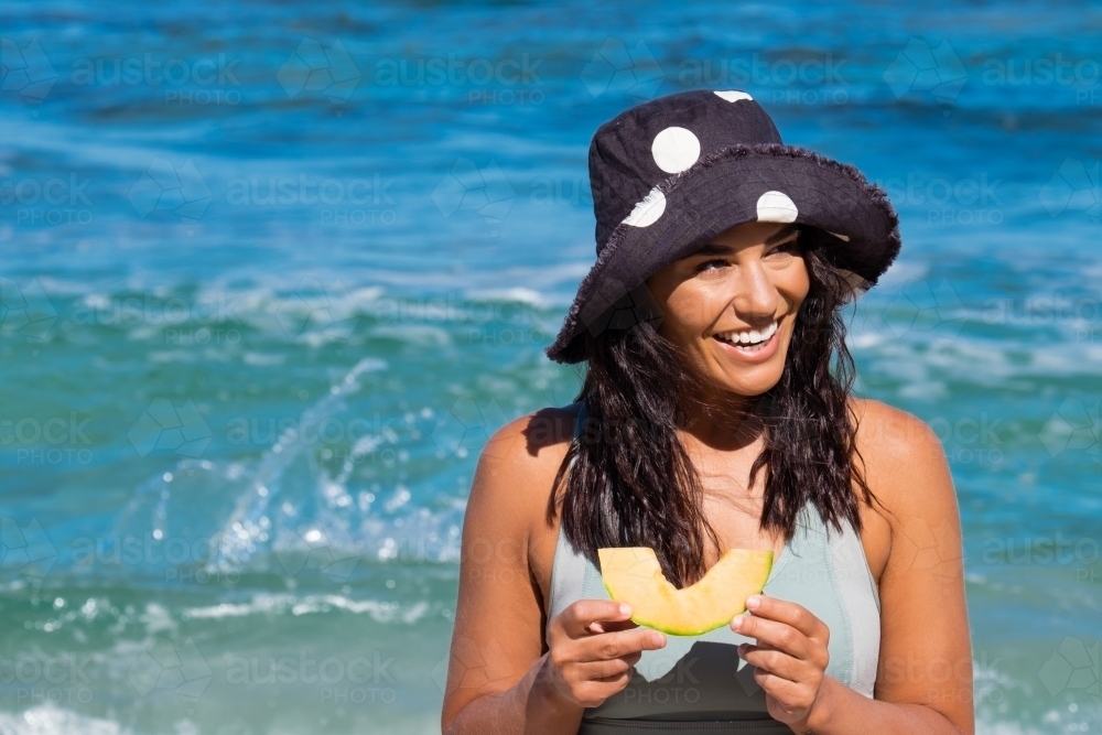 Image of Close up of Young woman wearing full brim hat and eating rockmelon  on the beach on clear sunny day - Austockphoto
