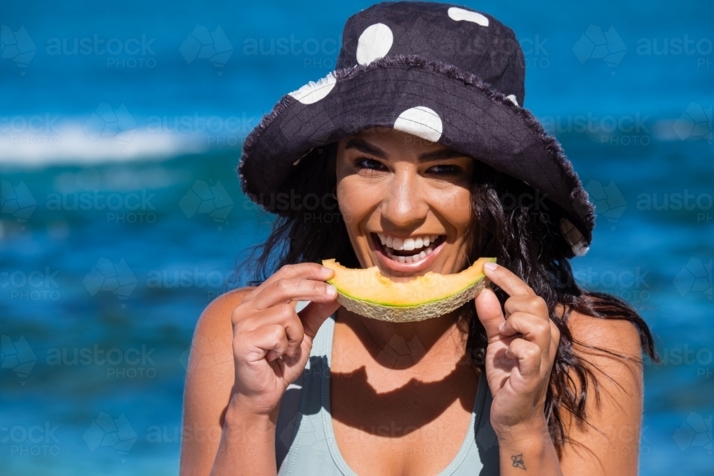 Close up of Young woman wearing full brim hat and eating rockmelon on the beach on clear sunny day - Australian Stock Image