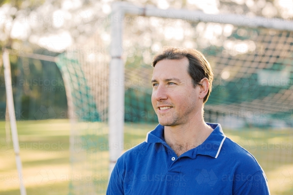 Close up photo of a smiling man wearing blue shirt on a sunny day with goal in the background - Australian Stock Image