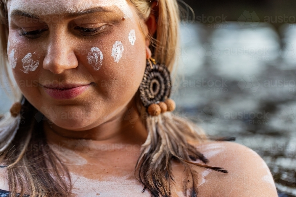 Close up portrait of aboriginal woman with woven earring and ochre paint on face looking down - Australian Stock Image