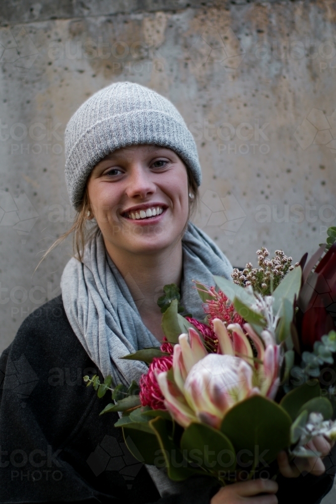 Close up portrait of young girl wearing a beanie and scarf smiling whilst holding a floral bouquet - Australian Stock Image