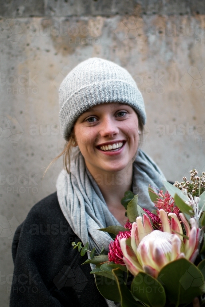 Close up portrait of young girl wearing a beanie and scarf smiling whilst holding a floral bouquet - Australian Stock Image