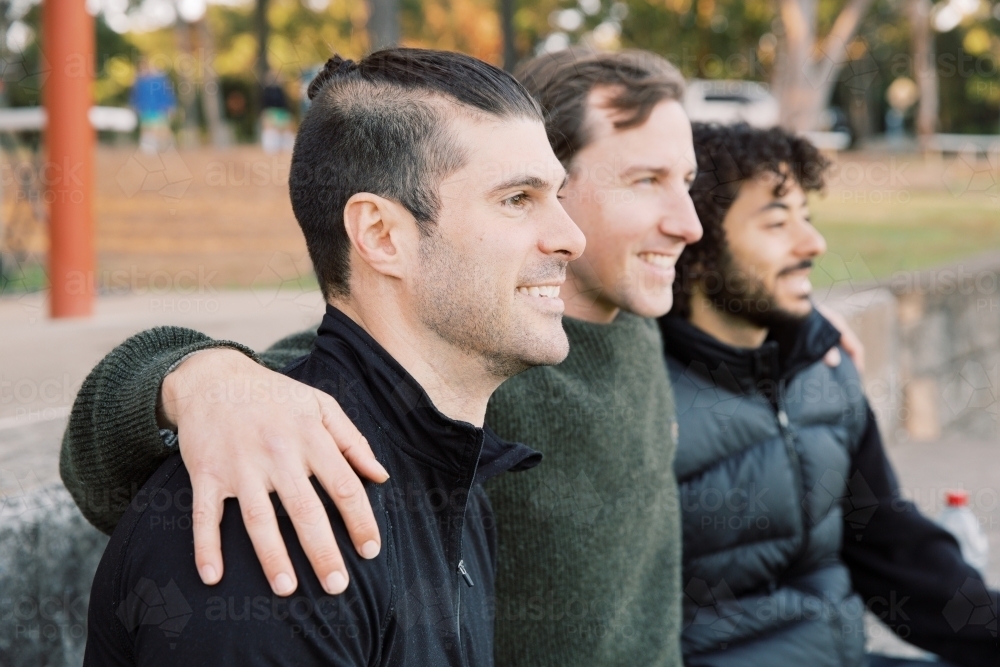 Close up shot of 3 smiling interracial men sitting on a field, hands on shoulders - Australian Stock Image