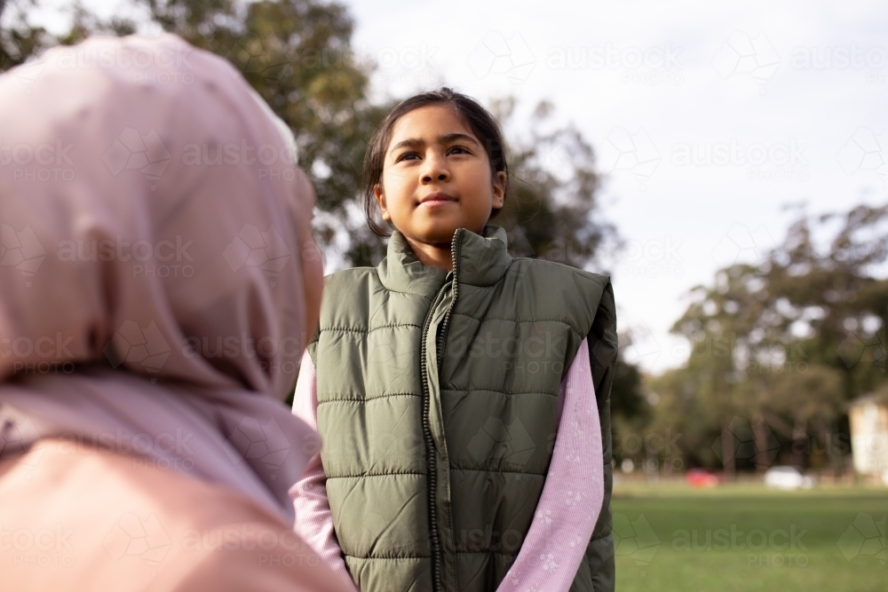 close up shot of a girl wearing green coat looking away with a woman wearing pink hijab in front - Australian Stock Image