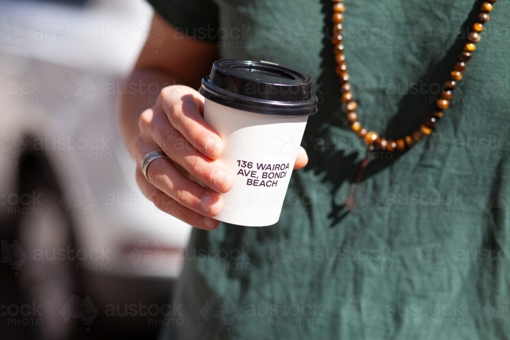 close up shot of a man holding a cup of coffee with wooden necklace and a ring and car at the back - Australian Stock Image