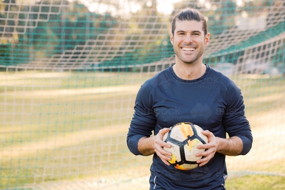 Close up shot of a smiling young man standing on the field , holding a soccer ball with a net behind - Australian Stock Image