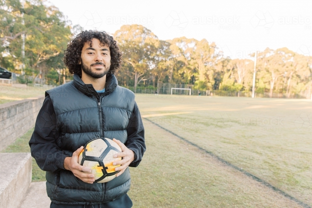 Close up shot of a smiling young man with curly hair standing on the field with a soccer ball - Australian Stock Image