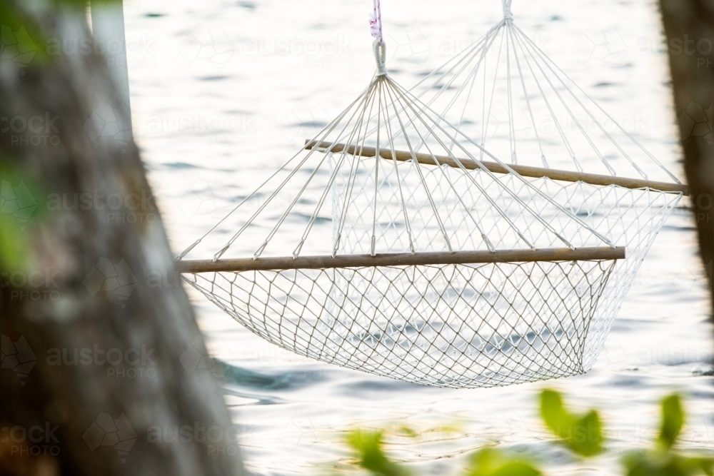 close up shot of a white string hammock near an ocean water with blurred green leaves - Australian Stock Image