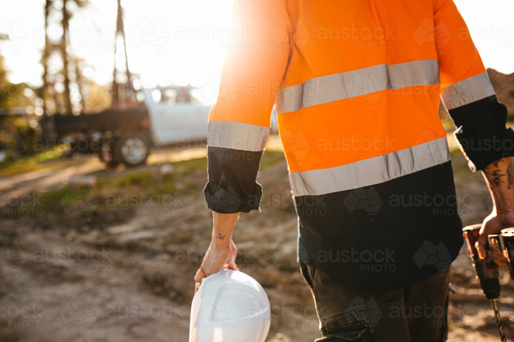 Construction worker holding a helmet and a drill while walking on the construction site. - Australian Stock Image