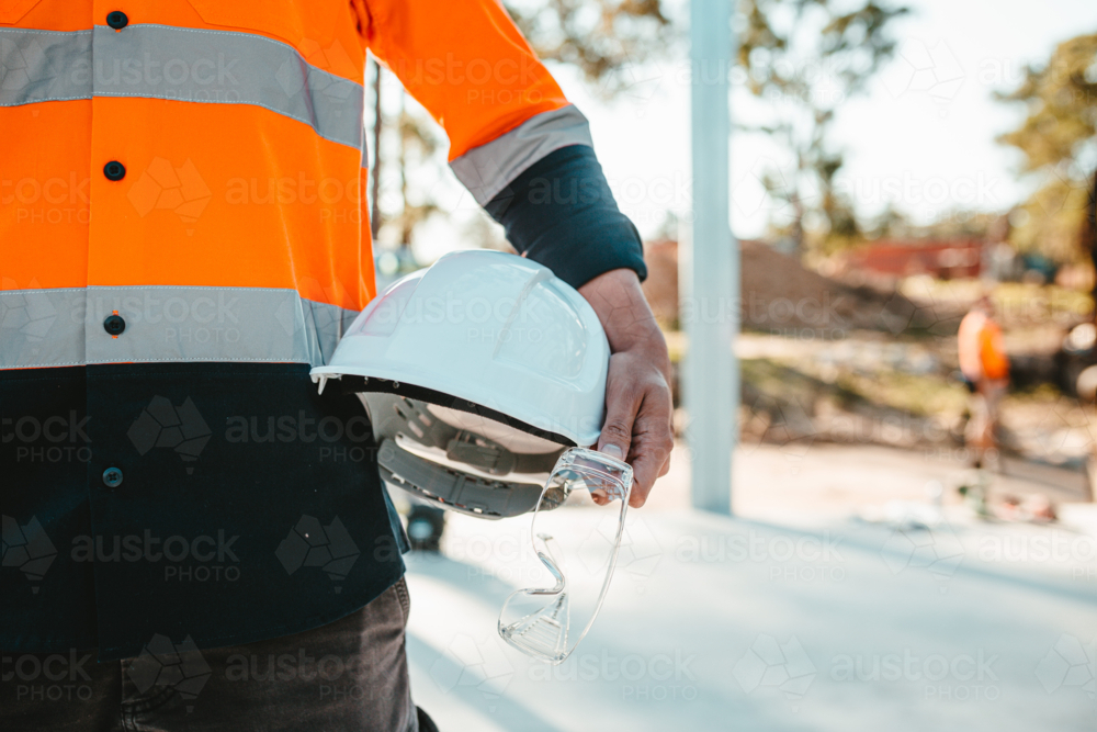 Construction worker holding a white helmet and safety glasses at construction site. - Australian Stock Image