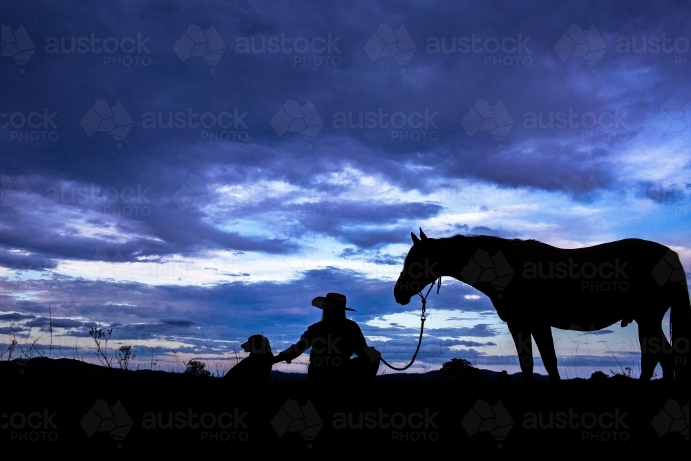 Country woman on farm crouched beside dog and horse silhouette - Australian Stock Image