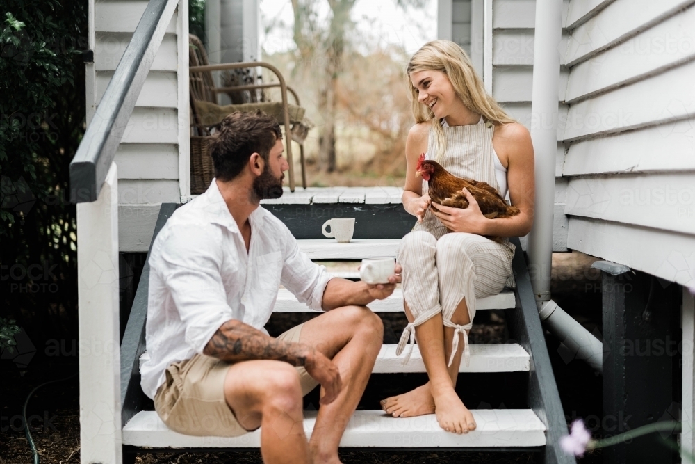 Couple having a cup of coffee on steps of verandah, with woman holding a chicken. - Australian Stock Image