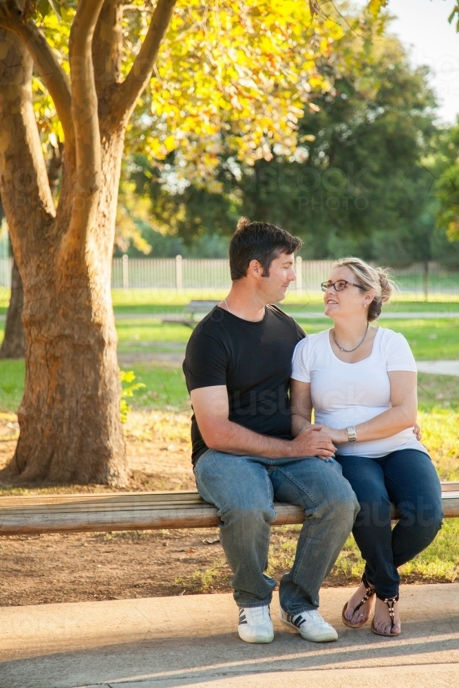 Couple sitting on park bench under autumn tree looking at each other - Australian Stock Image