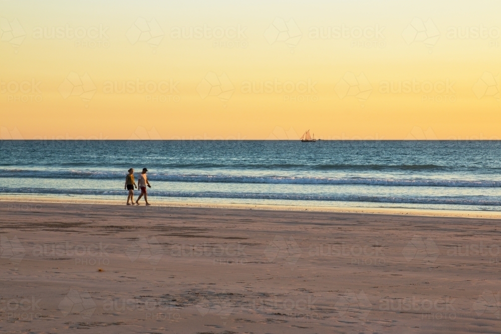 Couple walking along beach at sunset with boat sailing by - Australian Stock Image