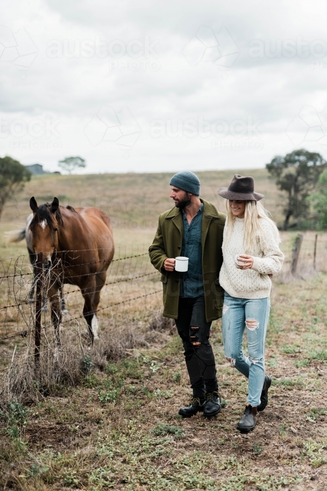 Couple walking past horses on a farm, holding cups of coffee - Australian Stock Image