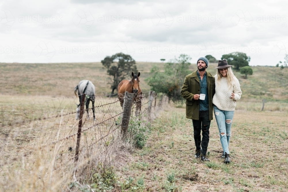 Couple walking past horses on a farm, holding cups of coffee - Australian Stock Image