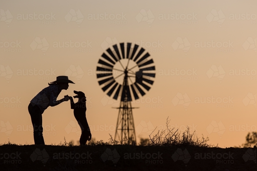 Cowgirl playing with dog silhouette with windmill - Australian Stock Image