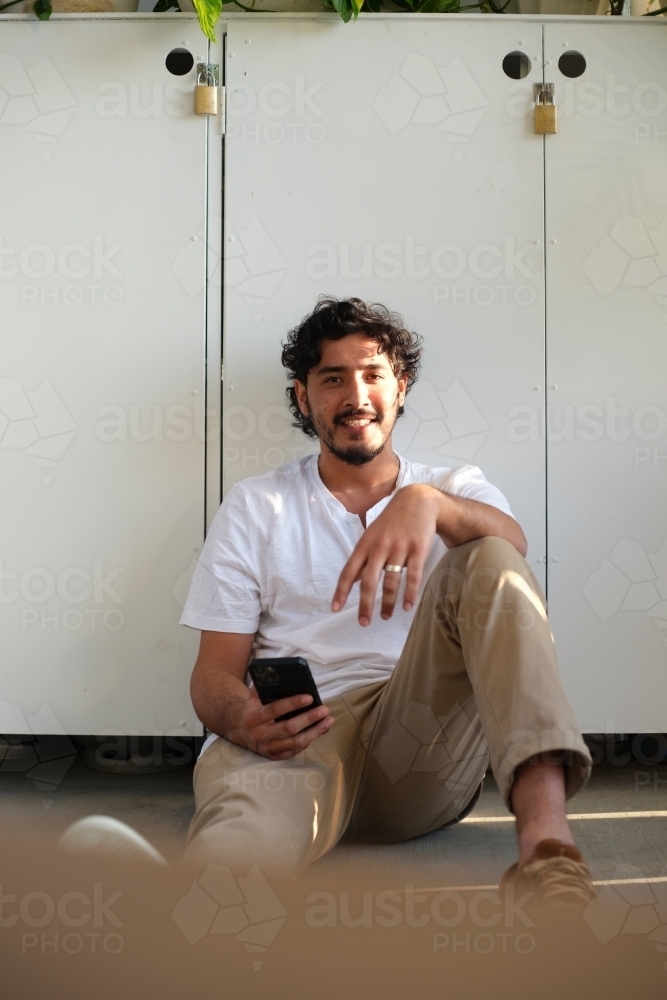 Curly haired man sitting on the ground while leaning on a cabinet while holding a mobile phone - Australian Stock Image