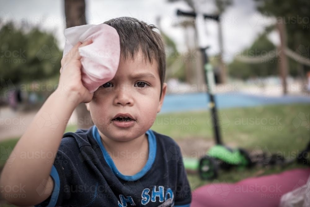 Cute 3 year old mixed race boy holds an esky ice pack on his head after falling off his scooter - Australian Stock Image