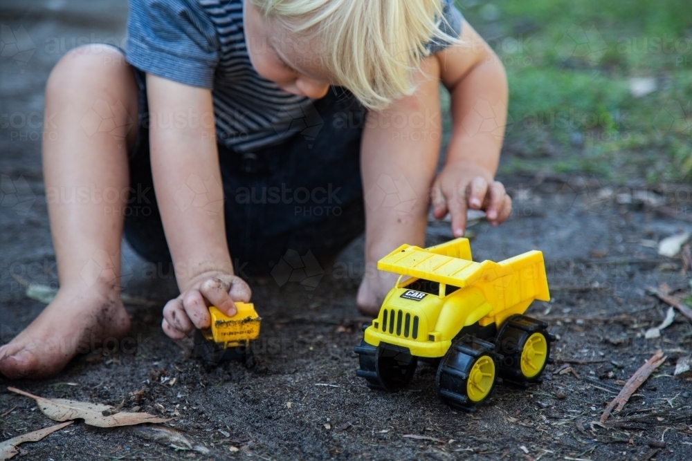 Cute little boy happily playing in black dirt with yellow toy trucks - Australian Stock Image