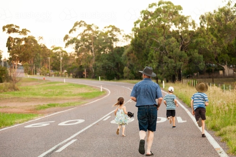 Dad and children running down country road in the evening - Australian Stock Image