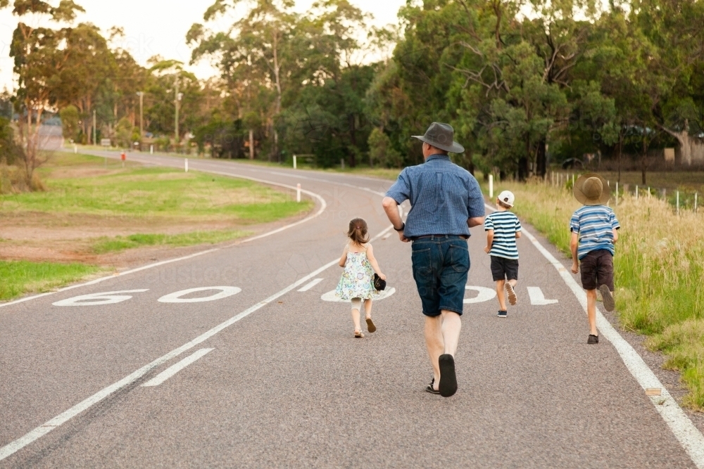 Dad and children running down country road in the evening - Australian Stock Image