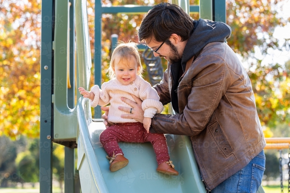 Dad helping happy baby girl slide down park slippery dip in autumn - Australian Stock Image