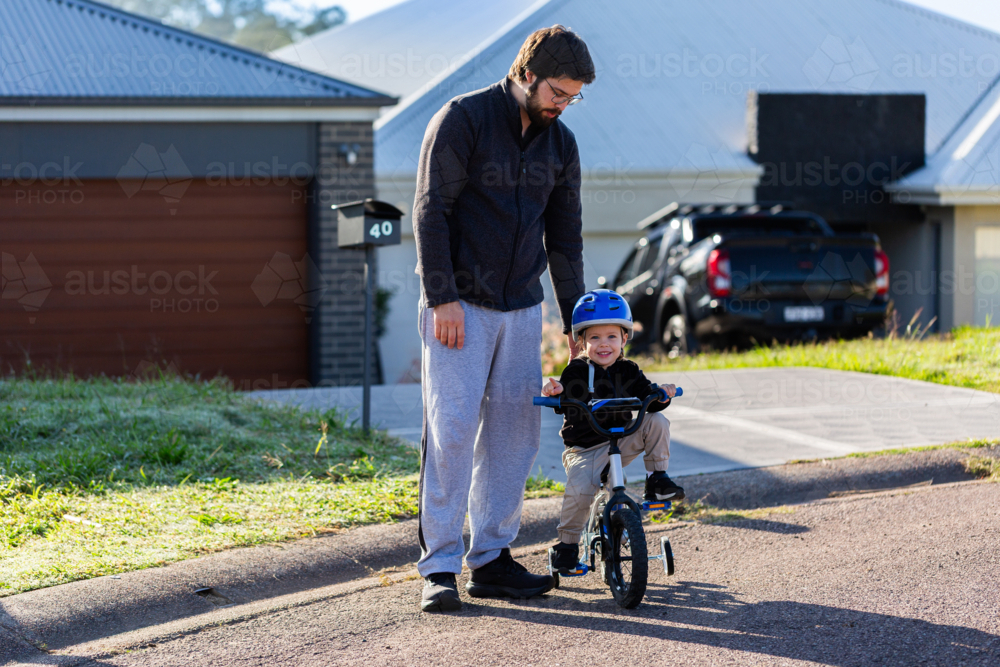 dad helping little girl learning to ride her bike with training wheels on quiet suburban street - Australian Stock Image