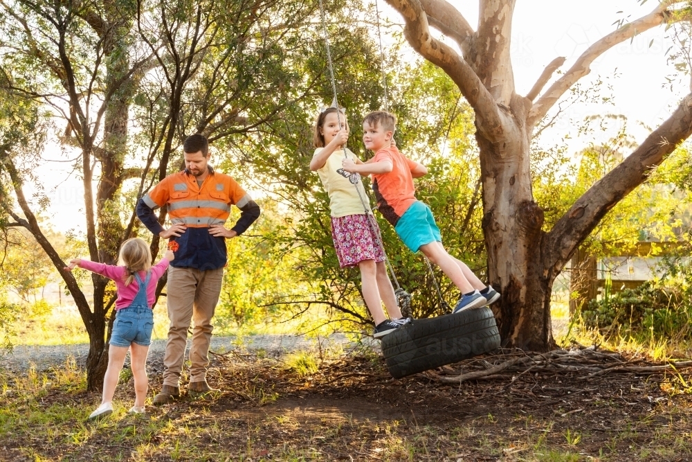 Dad playing with children in backyard on tyre swing in the afternoon - Australian Stock Image