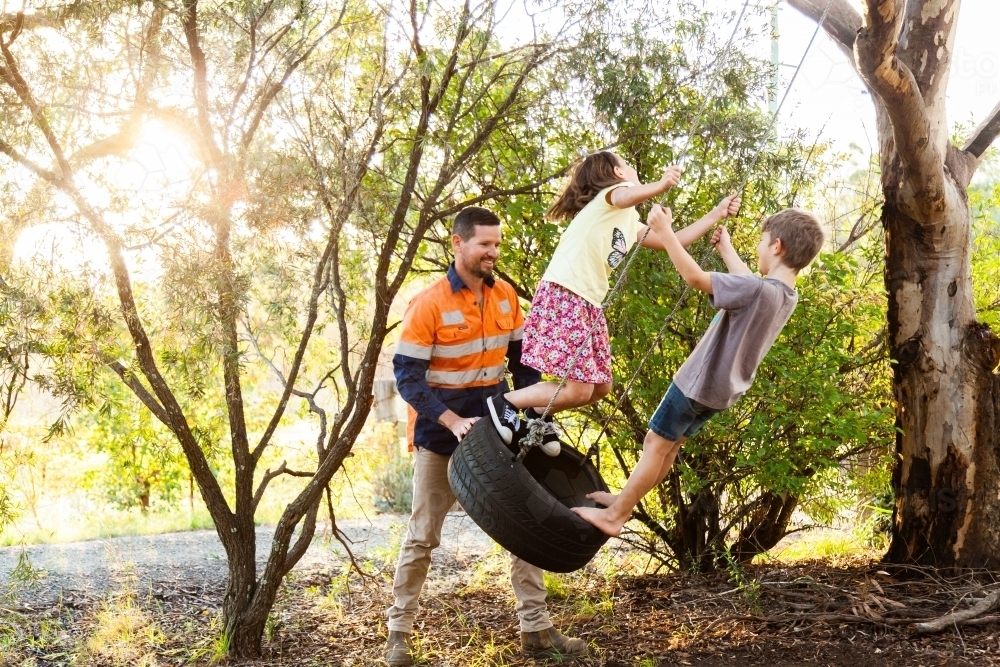 Dad pushing children on tyre swing boy and girl - Australian Stock Image