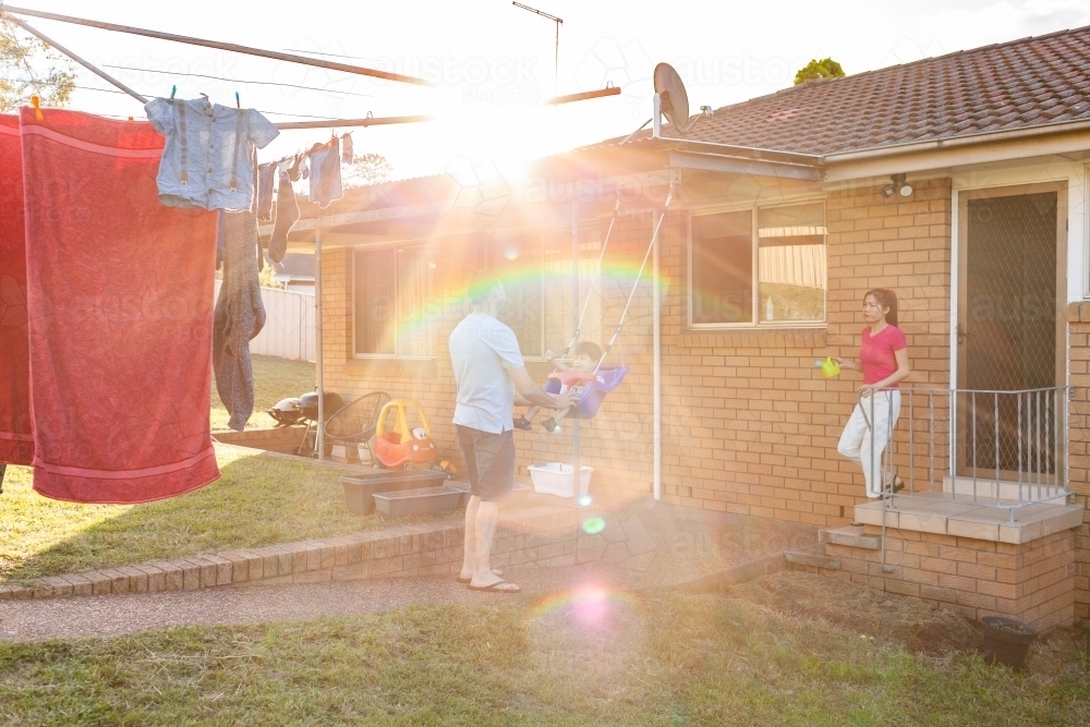 Dad pushing toddler boy on swing in backyard with sun flare - Australian Stock Image