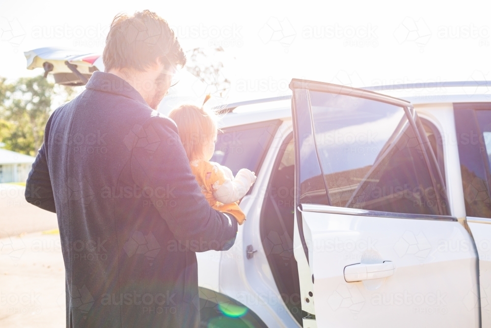 Dad putting baby girl in car with sun flare light - Australian Stock Image