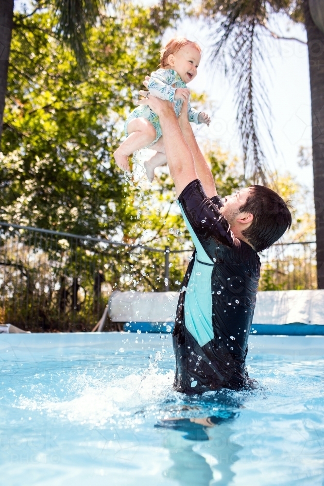 Dad throwing baby up in air in backyard pool during summer - Australian Stock Image