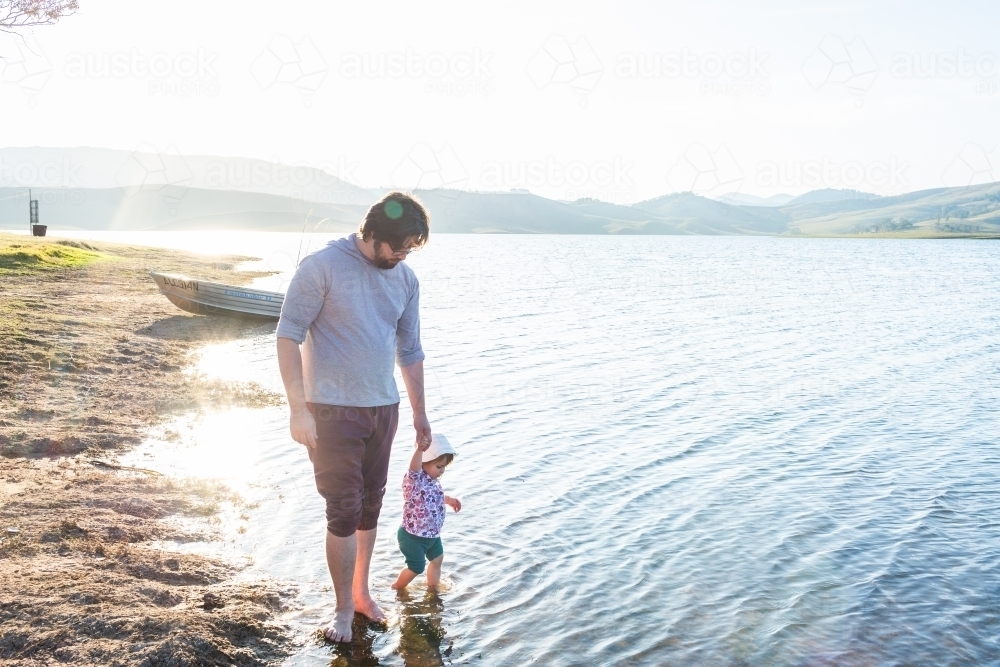 Dad with toddler wading in water by lakeside in afternoon together - Australian Stock Image
