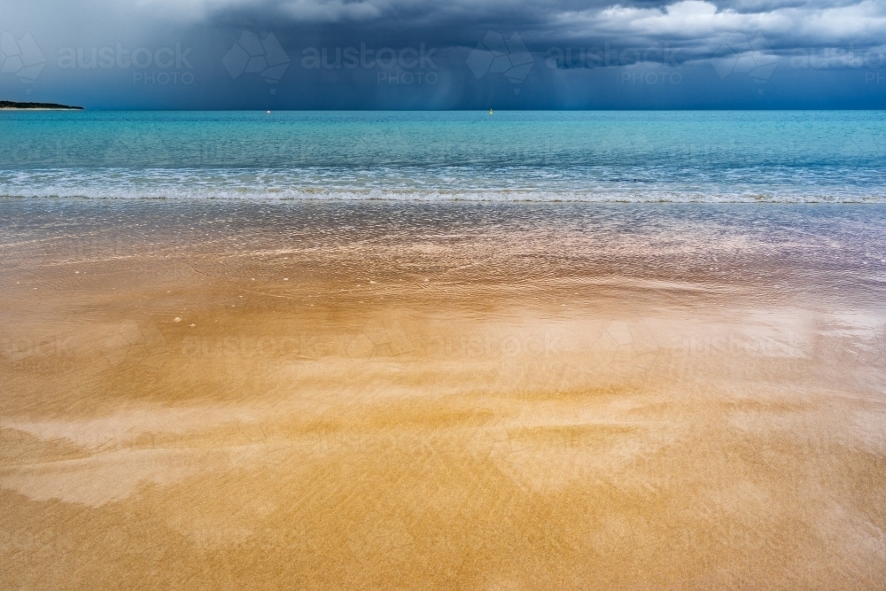 Dark storm clouds and rain over the ocean reflecting on a wet sandy beach - Australian Stock Image