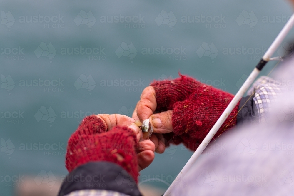 detail of hands of elderly person wearing mittens putting bait on hook while fishing - Australian Stock Image