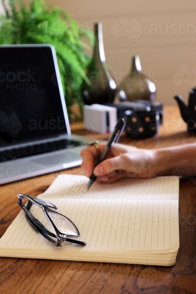 Detail shot of a hand writing in a note book with a laptop in background - Australian Stock Image